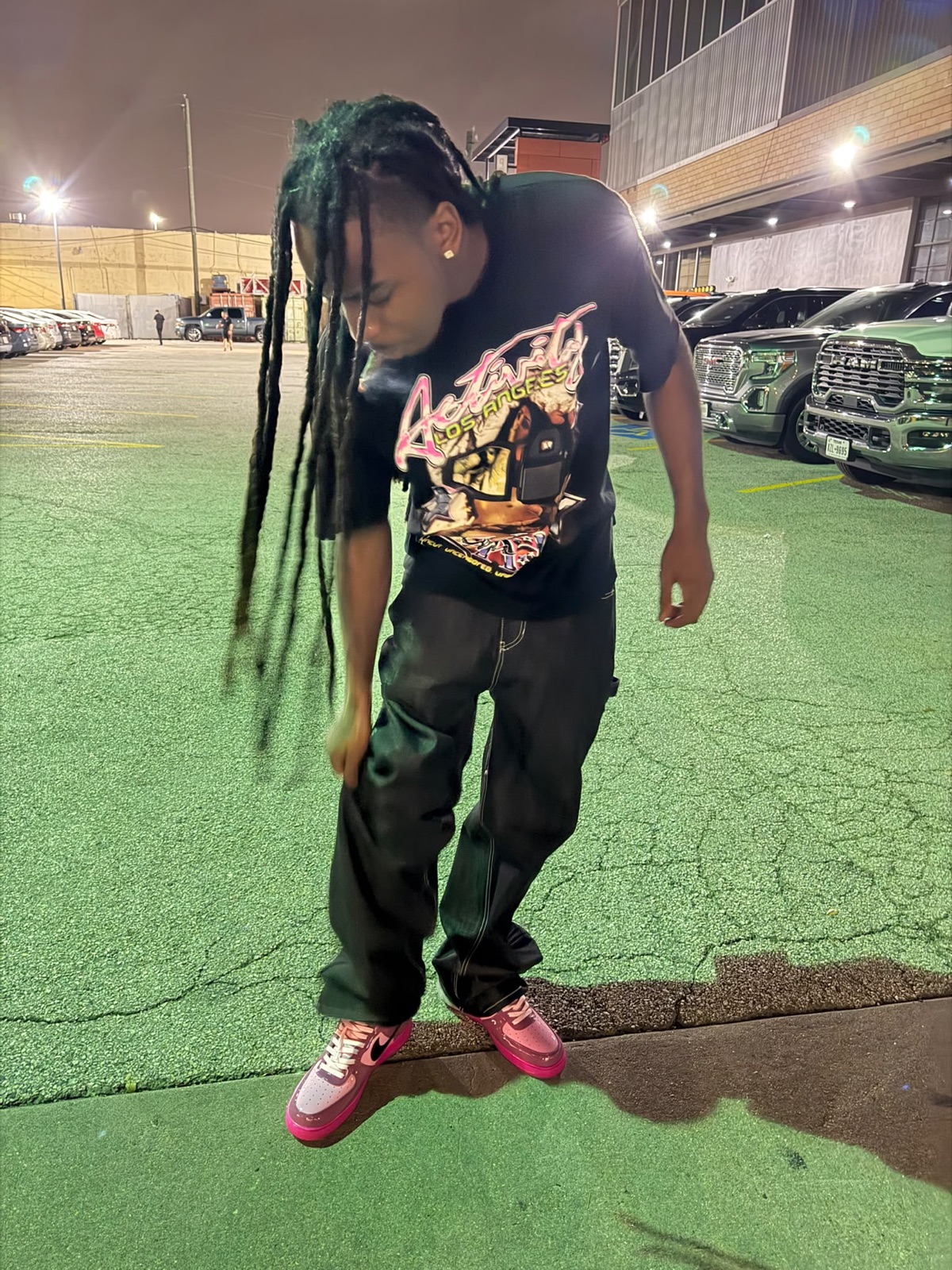 FMR Trill mid-motion in a night parking lot, dreads swinging, in a graphic tee and pink sneakers